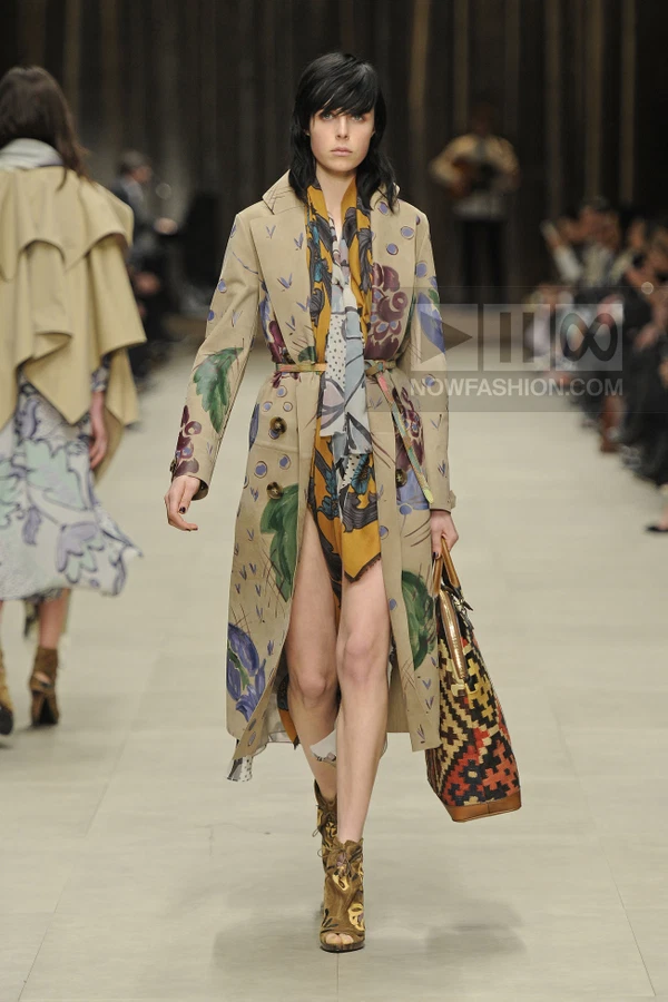 Model walking in a beige trench coat with hand-painted floral motifs, a patterned scarf, and a woven tote bag.