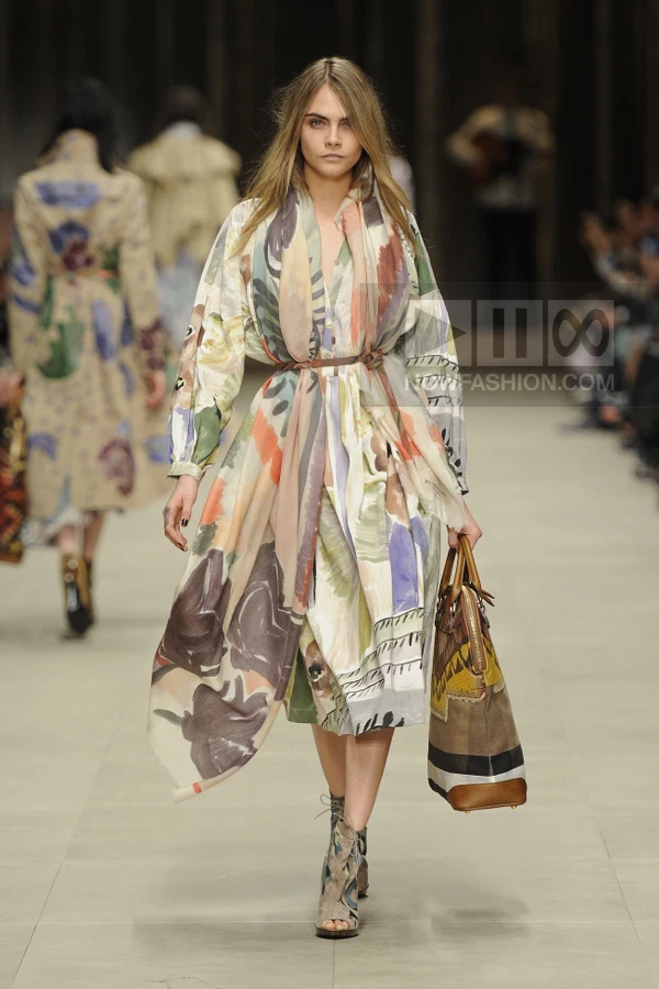 Model wearing a printed midi dress with a thin brown waist belt, carrying a large patterned tote bag on the runway.