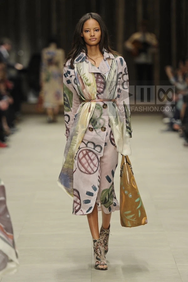 Model walking in a light pink trench coat with hand-painted abstract motifs, a matching scarf, and brown leather bag.