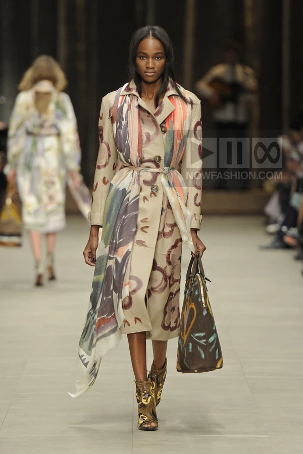 Model walking in a beige trench coat with hand-painted floral motifs, a long scarf, and a brown patterned handbag.