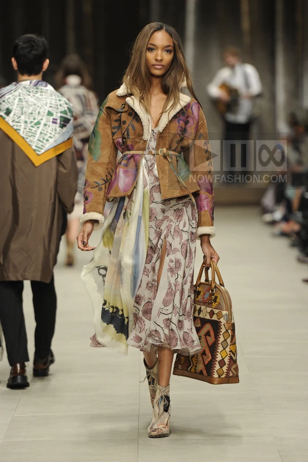 Model wearing a brown shearling-lined jacket over a floral midi dress, carrying a patterned tote bag on a runway.