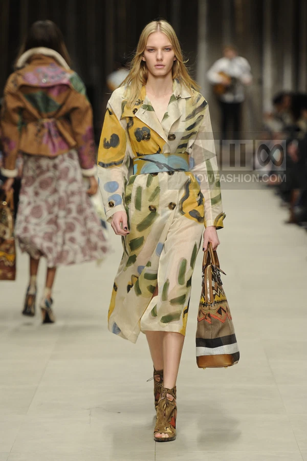 Model walking in a beige trench coat with hand-painted abstract brushstrokes, cinched with a wide blue painted belt.