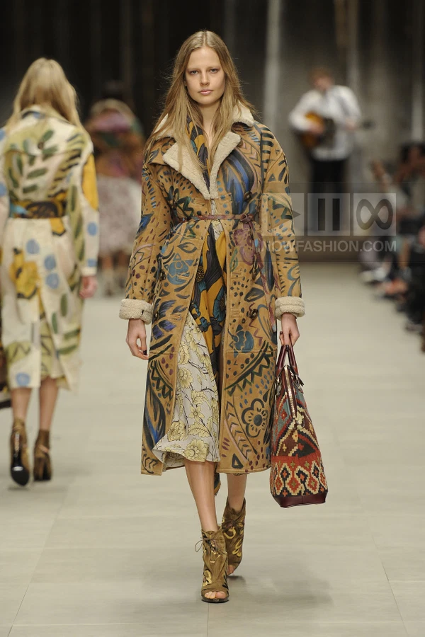 Model walking in a tan shearling-lined coat with hand-painted floral motifs, carrying a patterned tote bag on a runway.