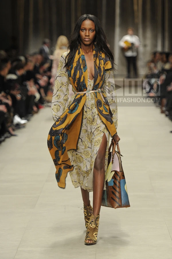 Model walking in a floral print midi dress with a draped mustard scarf, carrying a large patterned tote bag.