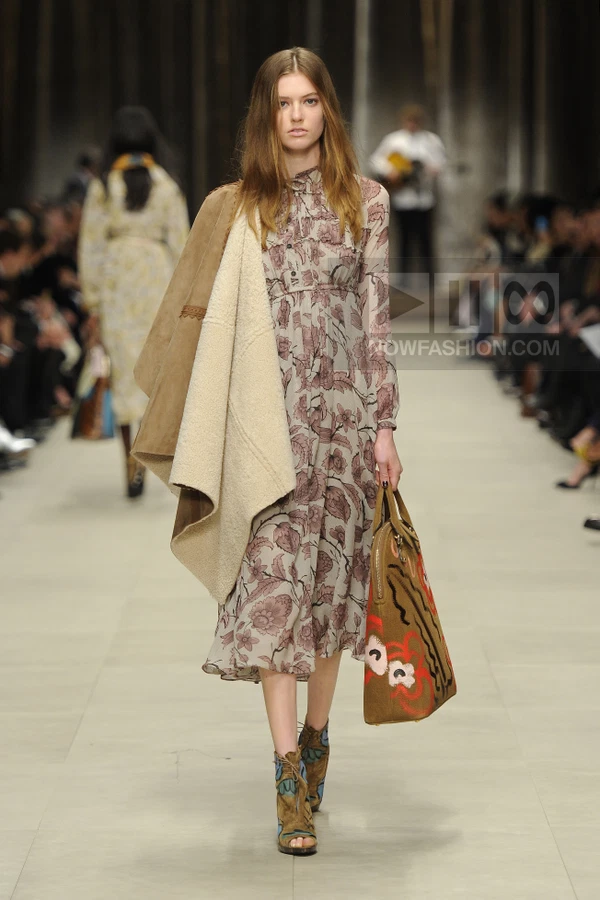 Model walking in a floral print midi dress with a shearling-lined suede coat draped over one shoulder, carrying a tote.