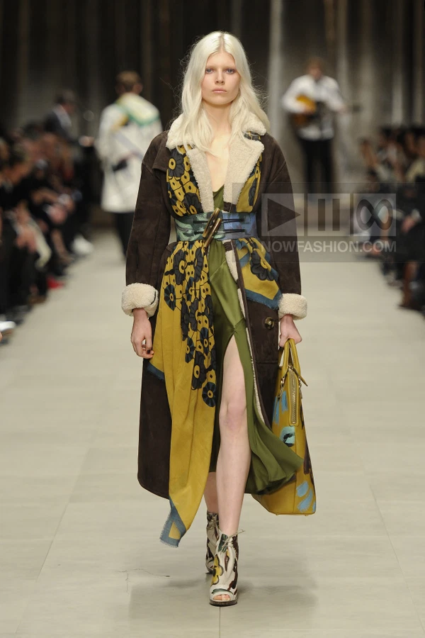 Model on runway wearing a brown suede coat with shearling trim over a green dress with a large floral print scarf.