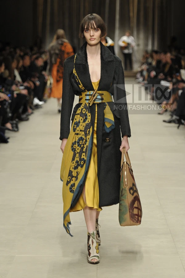 Model walking in a black wool coat over a yellow silk dress with a printed scarf tied at the waist and a painted tote.