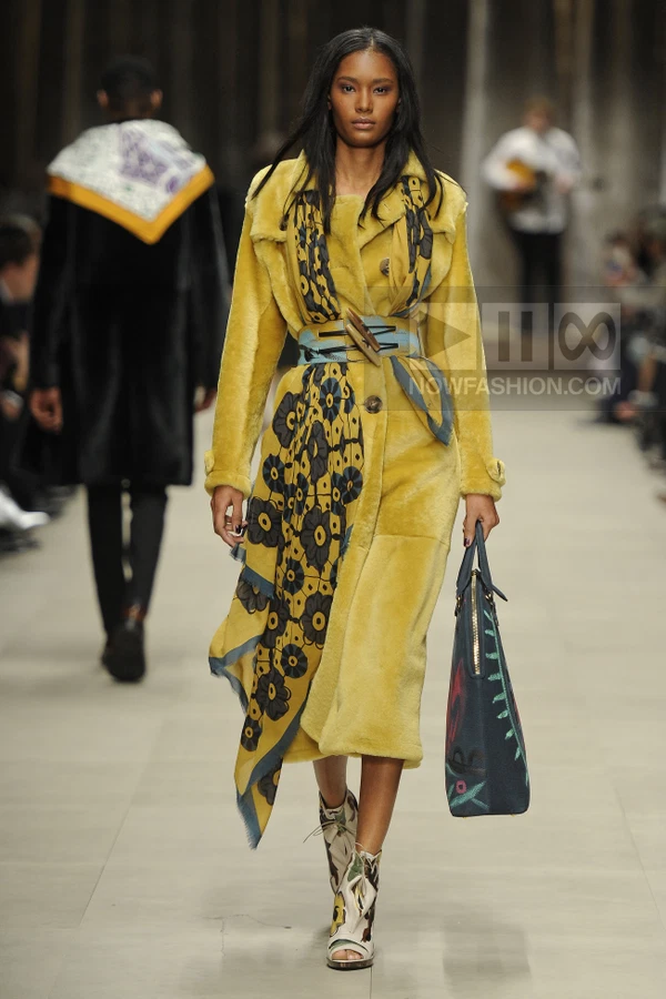 Model walking in a yellow shearling coat with a floral scarf, blue waist belt, and patterned open-toe ankle boots.
