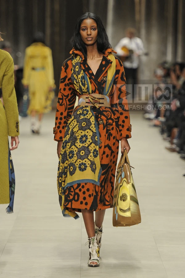 Model wearing an orange and black patterned wool coat with a floral scarf and wide leather belt, carrying a painted bag.