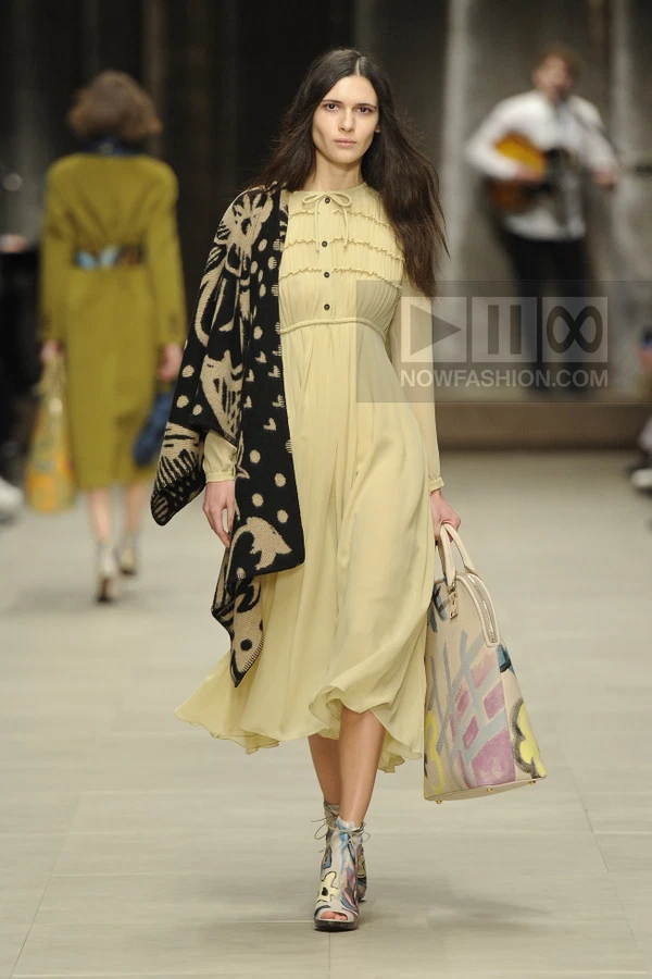 A model walks the runway wearing a pale yellow midi dress, a patterned black and beige shawl, and painted ankle boots.