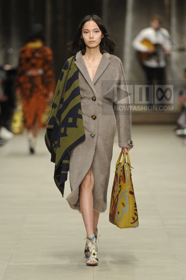 Model walking in a taupe mohair coat with a draped green and navy patterned shawl and a yellow printed tote bag.