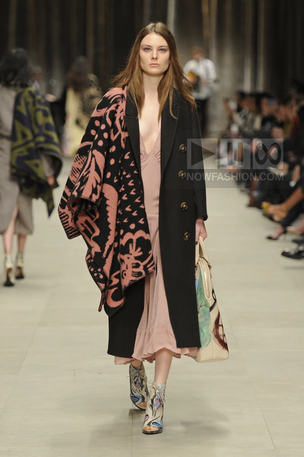 Model wearing a black wool coat over a blush pink slip dress, carrying a patterned tote bag on a runway.