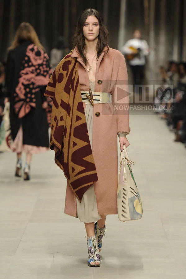 Model walking in a dusty pink mohair coat, beige midi dress, and patterned blanket scarf on a runway.