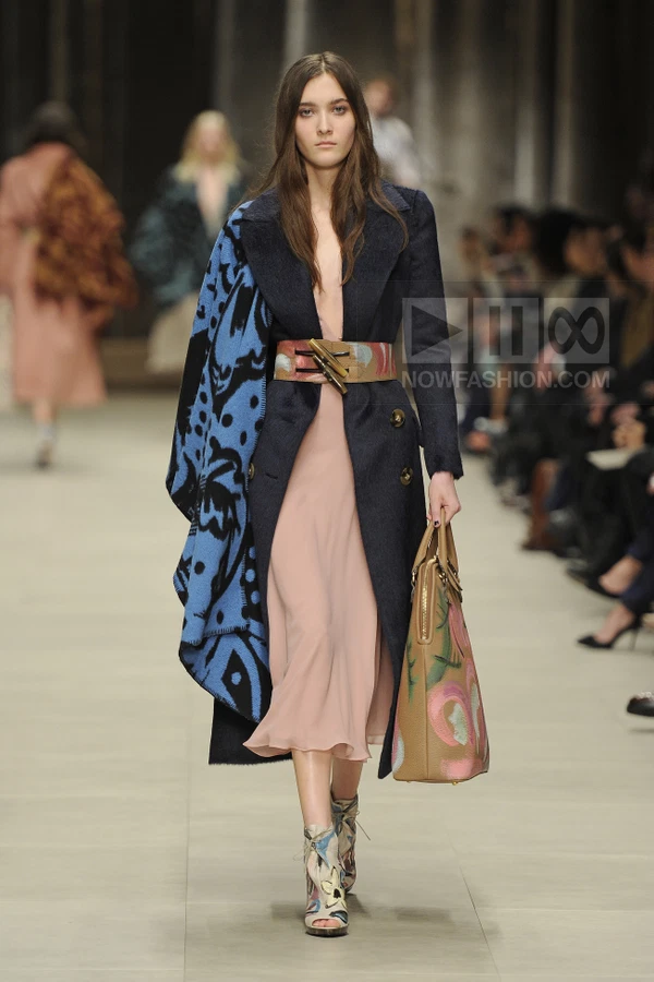 Model wearing a dark navy wool coat with a blue patterned blanket draped over one shoulder, over a pale pink midi dress.