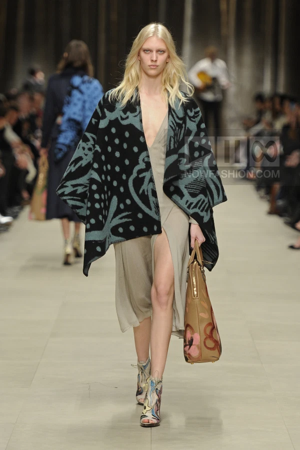 Model walking a runway wearing a beige midi dress, a patterned black and teal cape, and multi-colored heeled sandals.