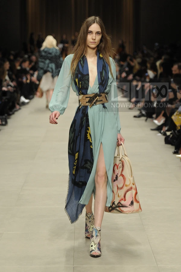 A model walks the runway in a light blue midi dress with a floral scarf, wide leather belt, and painted tote bag.