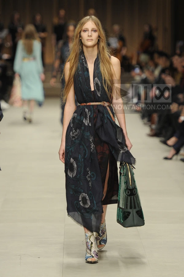 Model walking in a dark floral print scarf-dress with a leather belt and patterned open-toe booties on a runway.