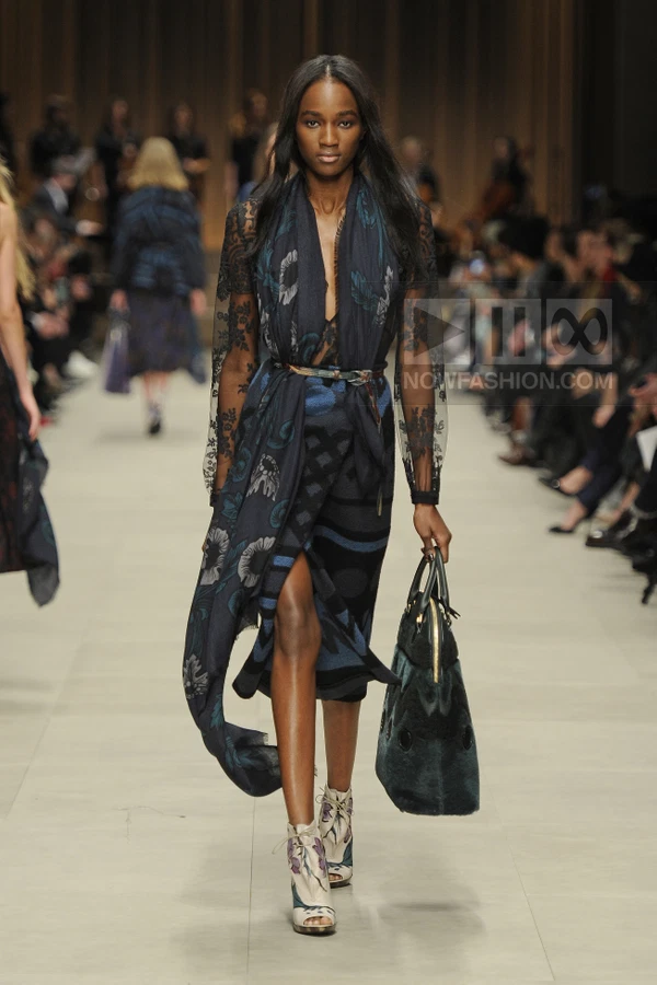 Model walking in a dark blue floral wrap dress with lace sleeves, a scarf, and a dark green fur-trimmed bag.