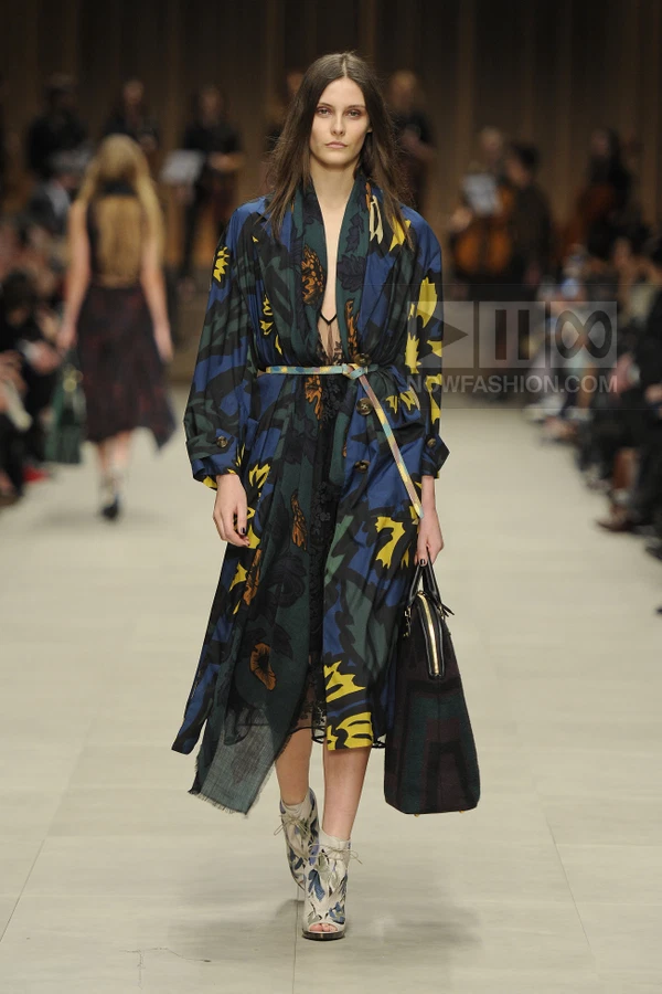 Model walking in a floral print trench coat over a black lace dress, carrying a large bag and wearing patterned boots.