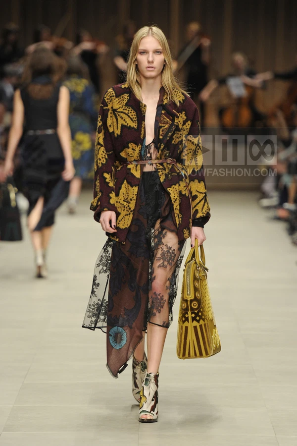 Model walking in a burgundy and yellow floral jacket, sheer lace skirt, and patterned ankle boots carrying a yellow bag.