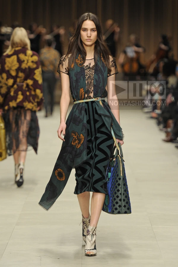 Model walking in a dark green floral scarf, lace top, and chevron velvet midi skirt, carrying a blue patterned bag.