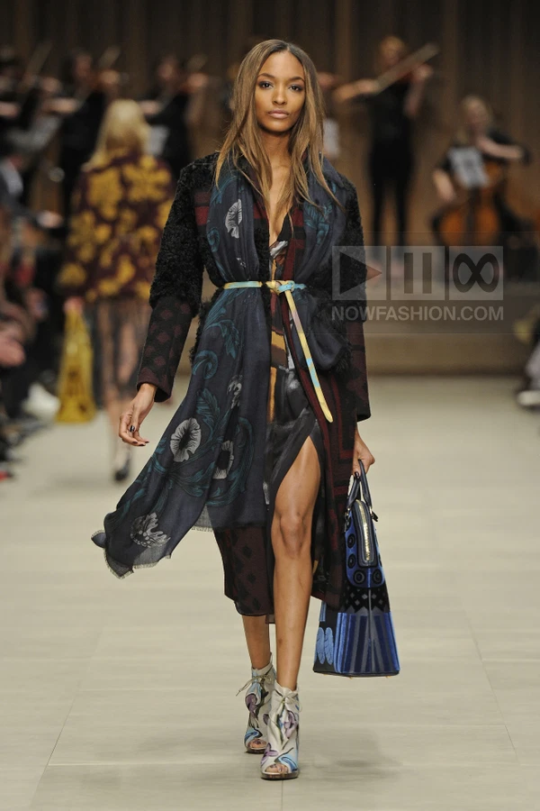 A model walks the runway wearing a patterned coat, a floral scarf, a belted dress, and printed open-toe booties.