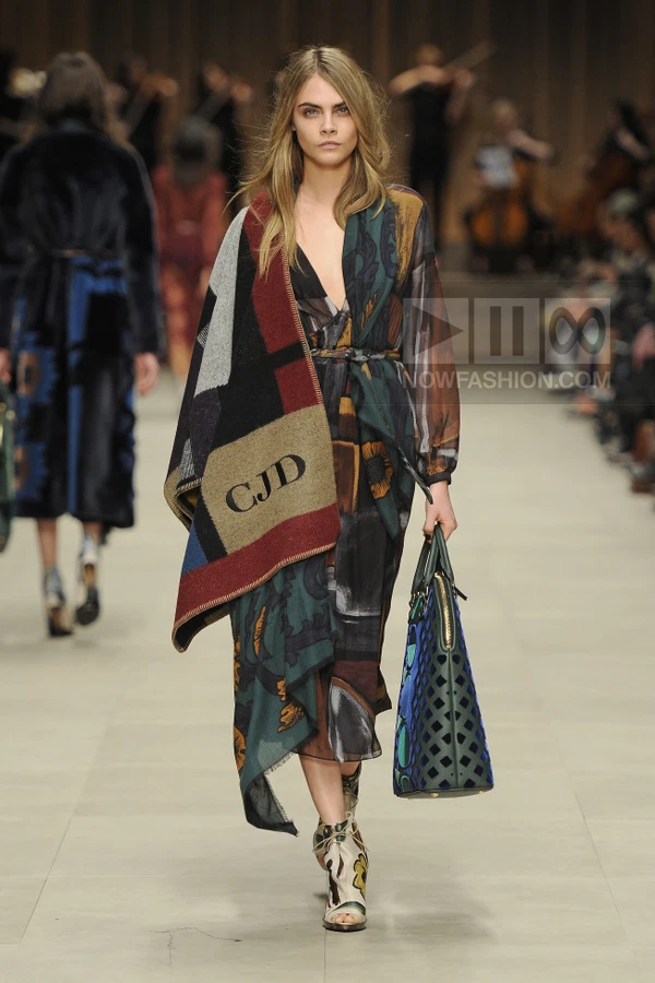 Model walking in a printed wrap dress, monogrammed blanket scarf, and laser-cut tote bag on a runway.