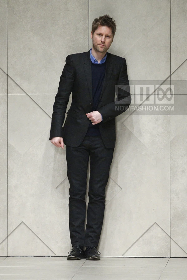 A person standing against a light gray geometric wall, wearing a black blazer, navy sweater, blue shirt, and black pants.