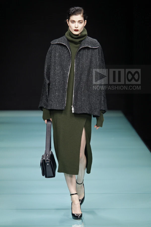 Model walking in a dark green ribbed knit midi dress with a side slit, layered under a charcoal textured zip-up jacket.