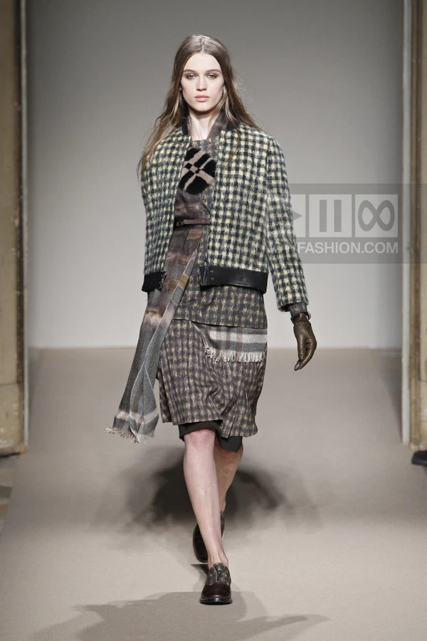 Model walking in a plaid jacket, layered plaid dress, scarf, leather gloves, and dark shoes on a runway.