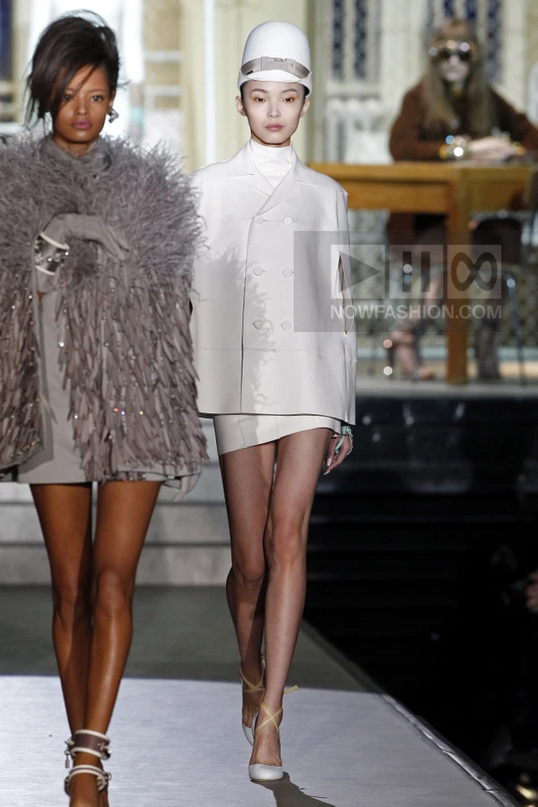Model in a white double-breasted cape jacket and matching mini skirt, wearing a white pillbox hat on a runway.
