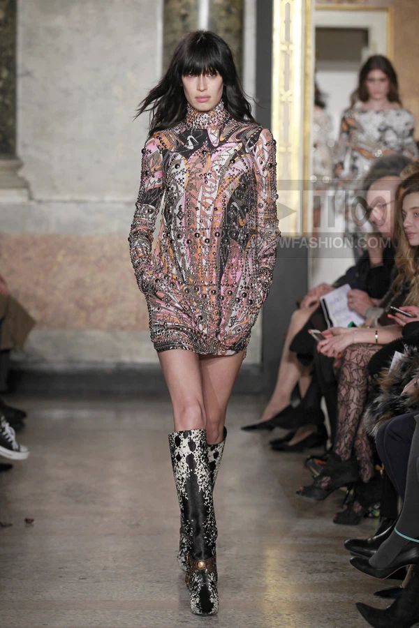 Model walking in a long-sleeve mini dress with a colorful abstract print and metal stud embellishments, paired with boots.