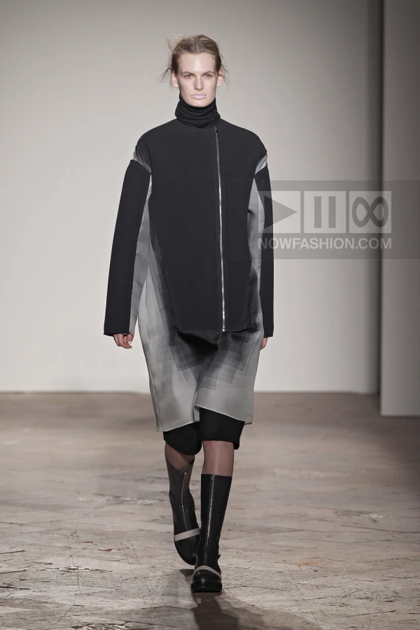 Model walking in a black and gray coat with a geometric print, black turtleneck, and black knee-high boots.