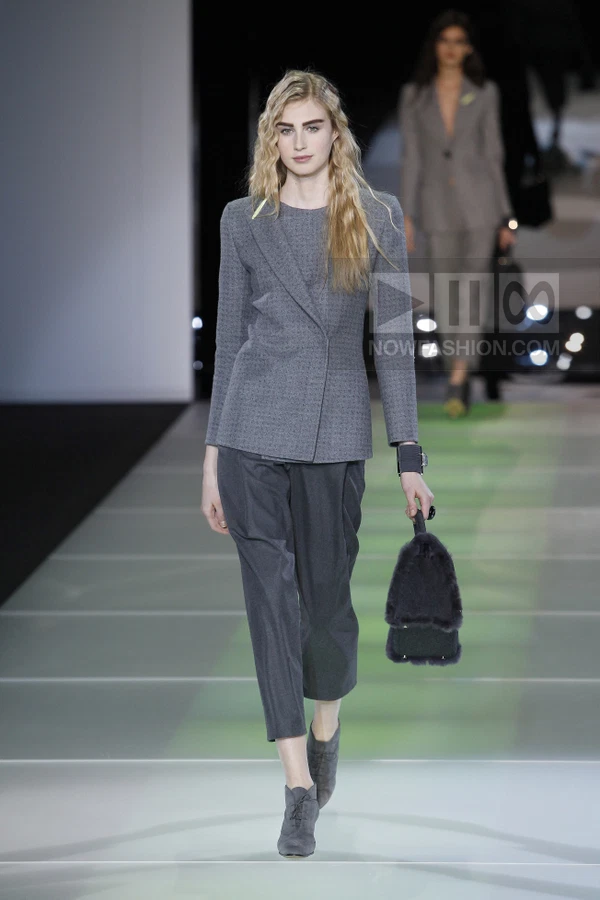 Model walking in a gray wool blazer, dark gray cropped trousers, suede lace-up shoes, and a small fur-trimmed handbag.