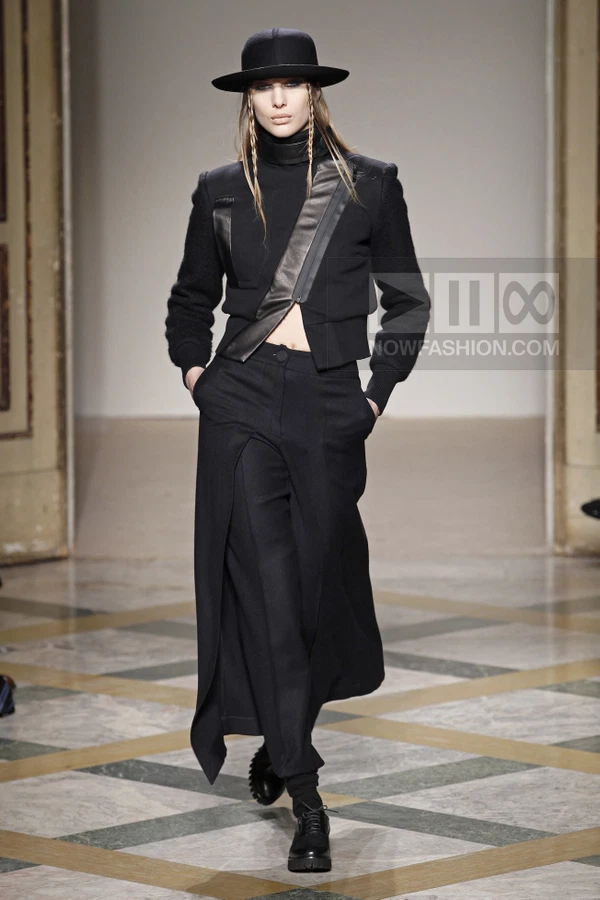 Model wearing a black cropped jacket, wide-leg trousers with an overskirt, and a fedora hat on a runway.