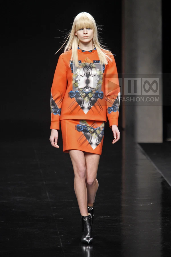 Model walking in an orange sweatshirt and matching mini skirt with a graphic face and rose print on a black runway.