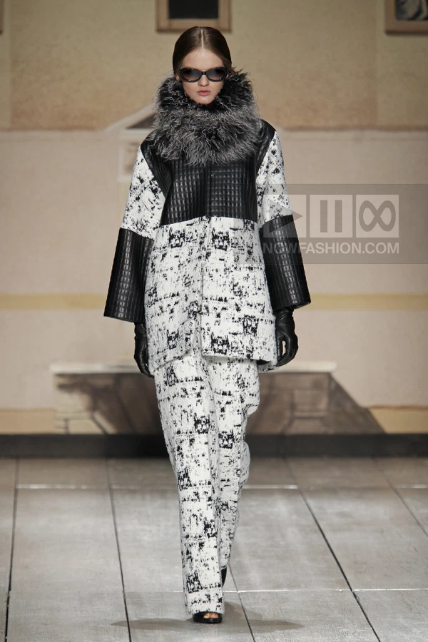 Model wearing a black and white patterned coat with a fur collar and matching trousers on a runway.