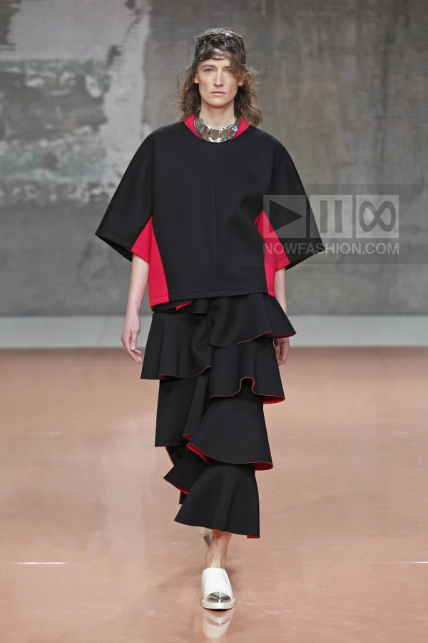 Model on runway wearing black top with red side panels and tiered ruffled skirt with red lining, silver collar necklace.