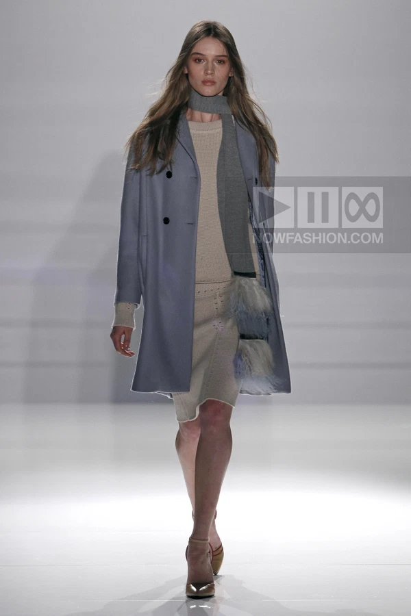 Model wearing a powder-blue double-breasted coat over a beige knit sweater and skirt with a gray scarf on the runway.