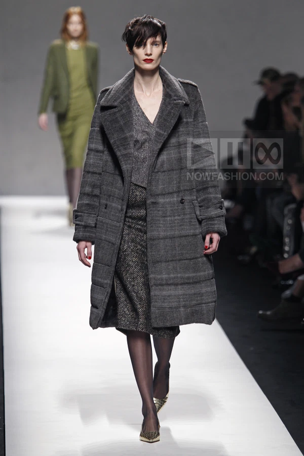 Model wearing a grey quilted coat over a dark herringbone skirt and metallic pointed-toe pumps on a runway.