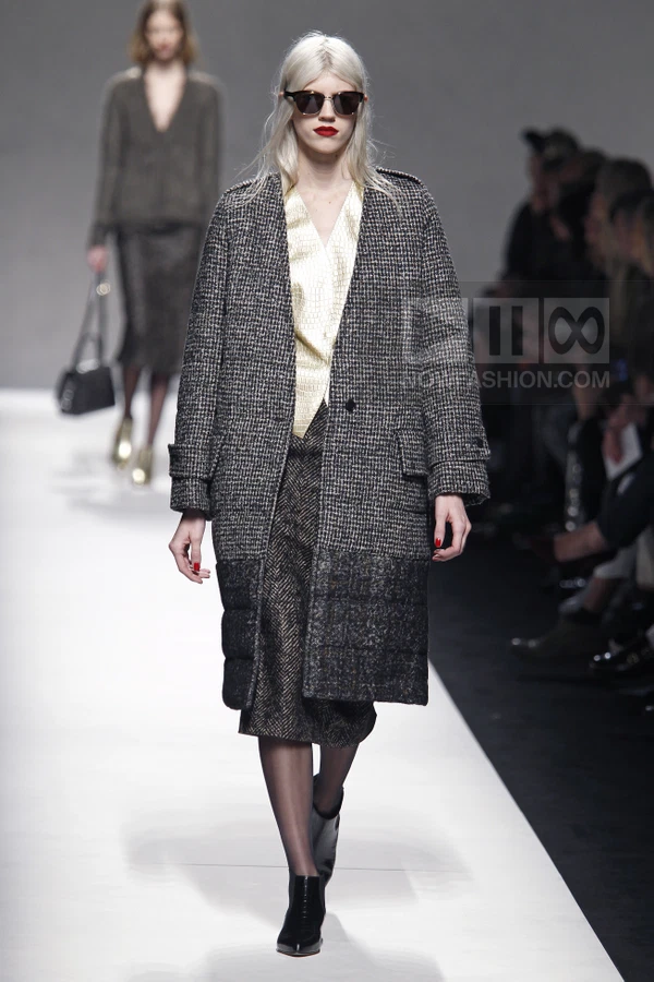 Model walking in a gray tweed coat with a quilted hem, a cream blouse, and a dark herringbone skirt on a runway.
