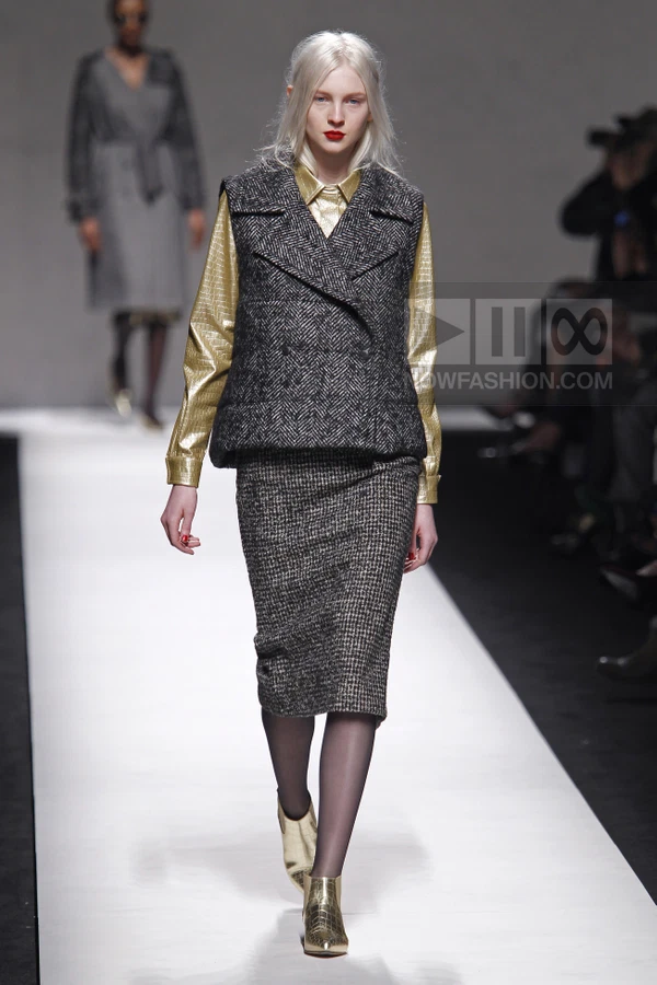Model walking in a metallic gold shirt, herringbone vest, and tweed pencil skirt on a white runway.