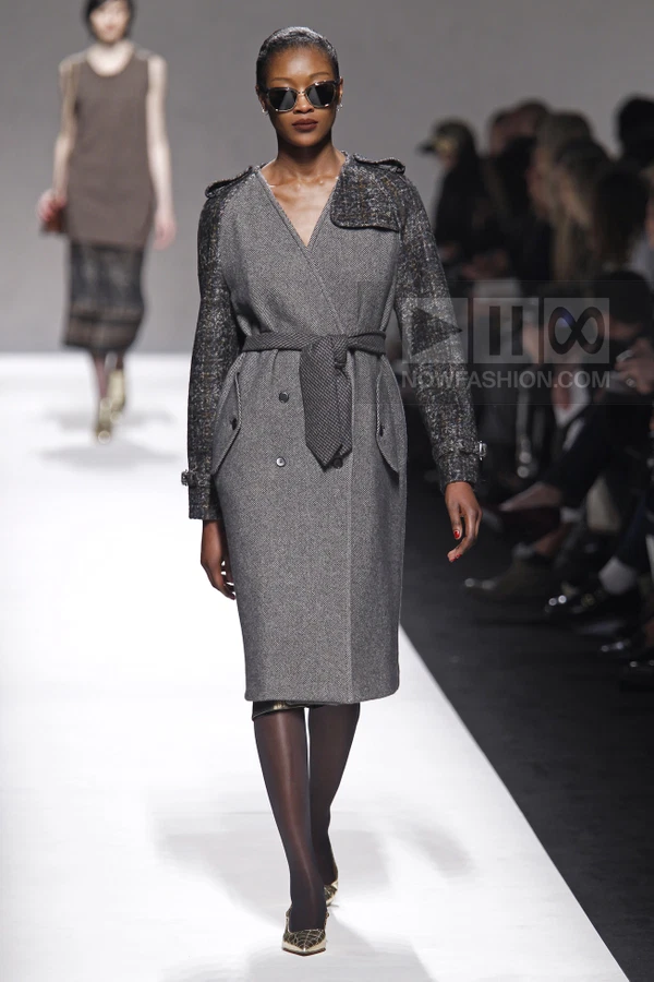 Model wearing a gray wool coat with dark textured sleeves and a belted waist, walking on a white runway.
