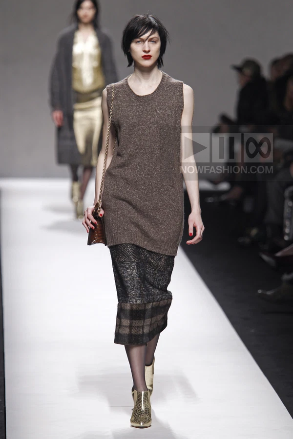 Model walking in a brown sleeveless knit tunic over a dark gray patterned midi skirt with gold ankle boots.