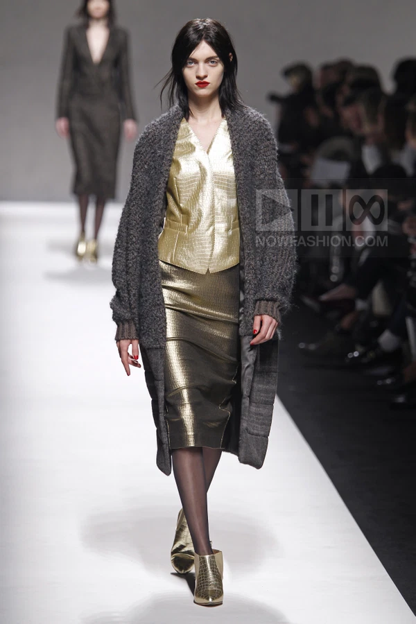 Model walking in a metallic gold vest and skirt, layered under a long, charcoal gray textured knit cardigan.