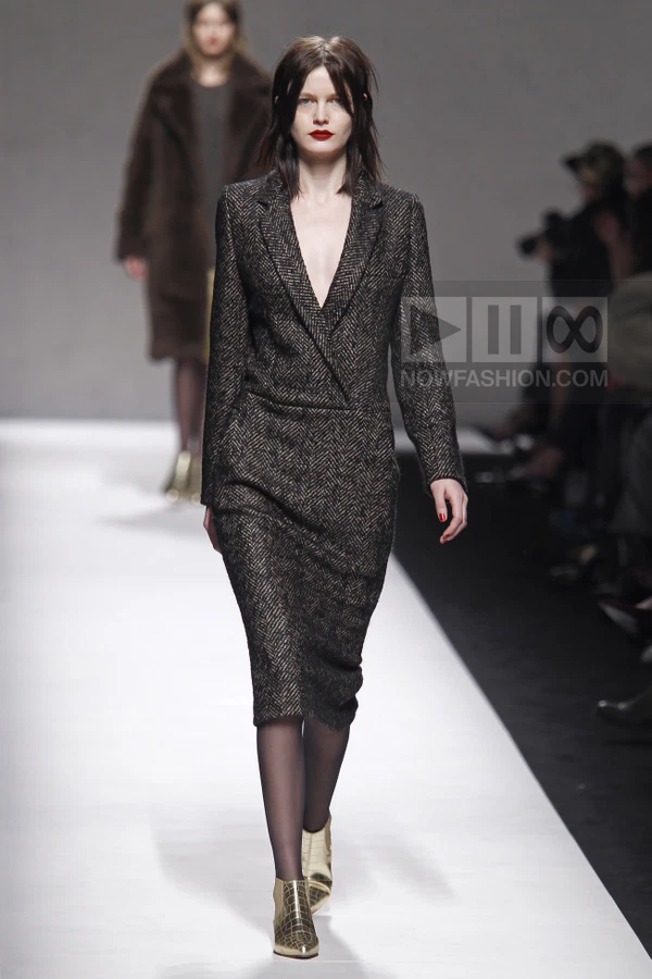 Model walking the runway in a dark brown herringbone wool blazer dress with a deep V-neckline and metallic gold boots.