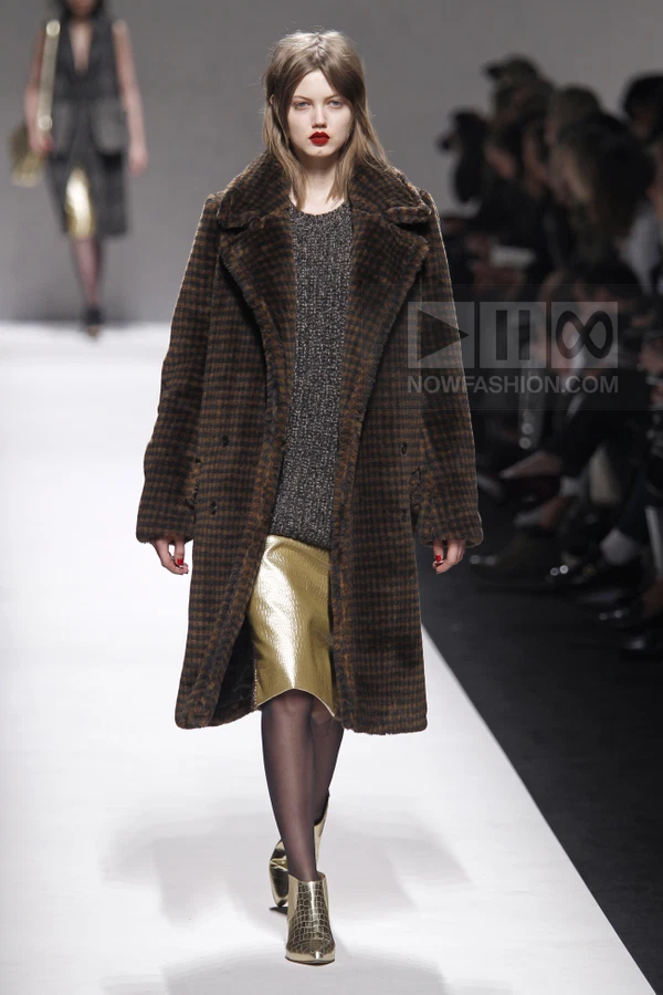 Model walking in a brown houndstooth coat over a gray knit sweater and a metallic gold skirt with gold ankle boots.