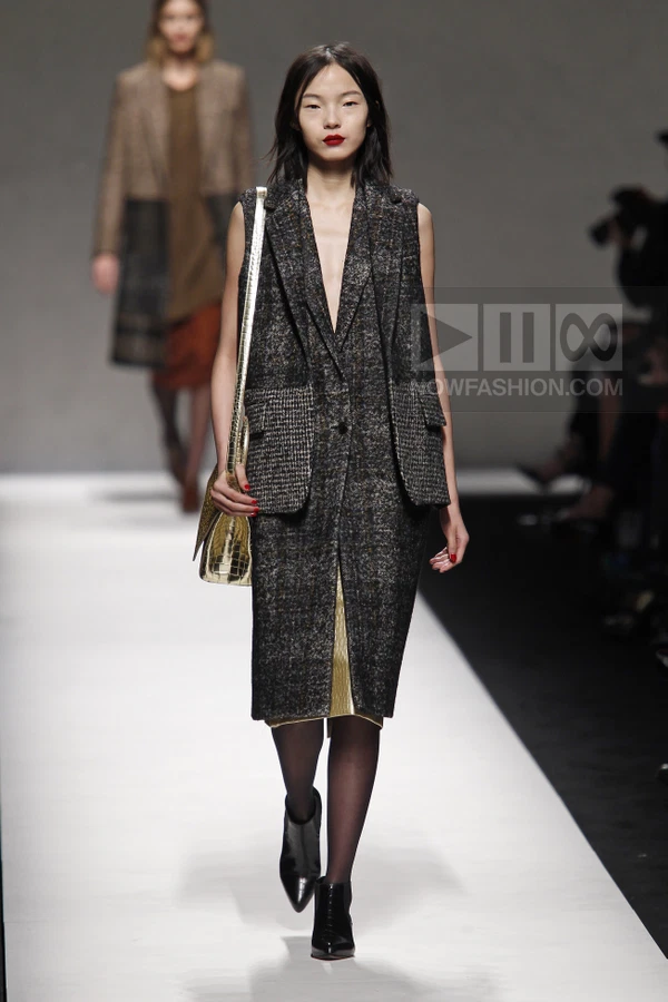 Model wearing a sleeveless tweed coat over a gold metallic skirt, with a gold shoulder bag and black ankle boots.