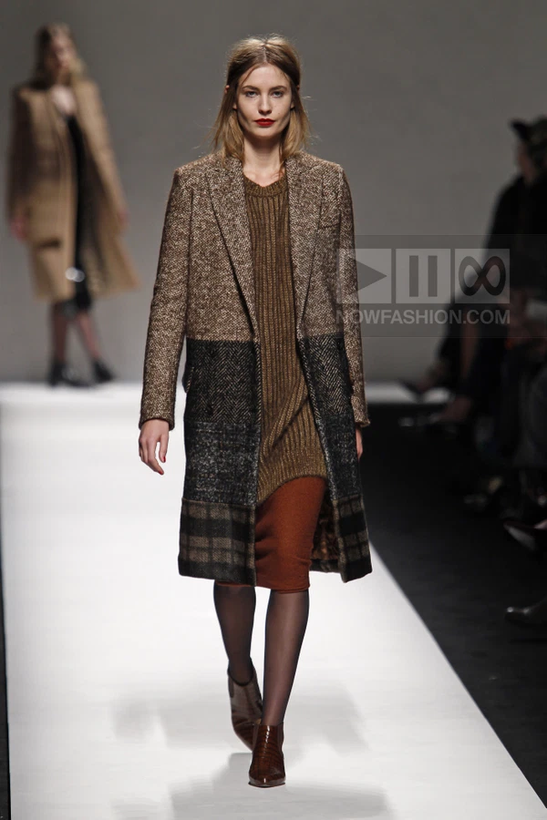 Model walking on a runway wearing a brown and black color-blocked wool coat over a ribbed knit dress and brown ankle boots.