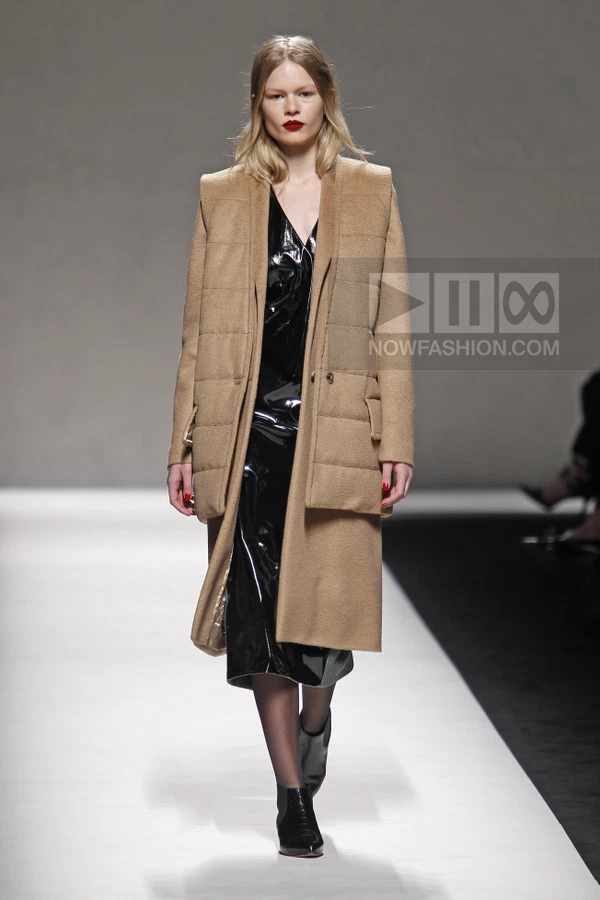 Model walking in a camel wool coat over a black patent leather midi dress with black ankle boots on a runway.